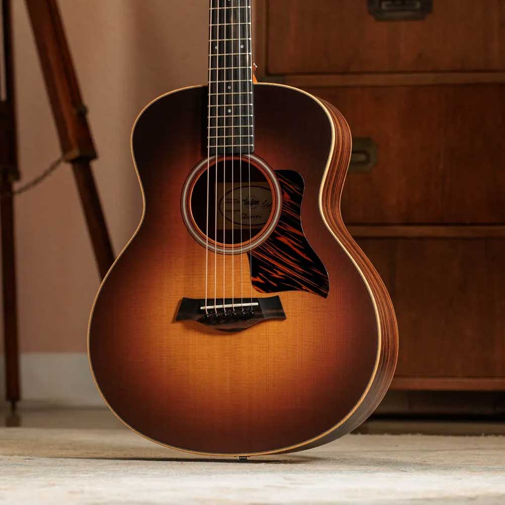Sunburst acoustic guitar resting on carpet indoors, with a wooden cabinet in the background.