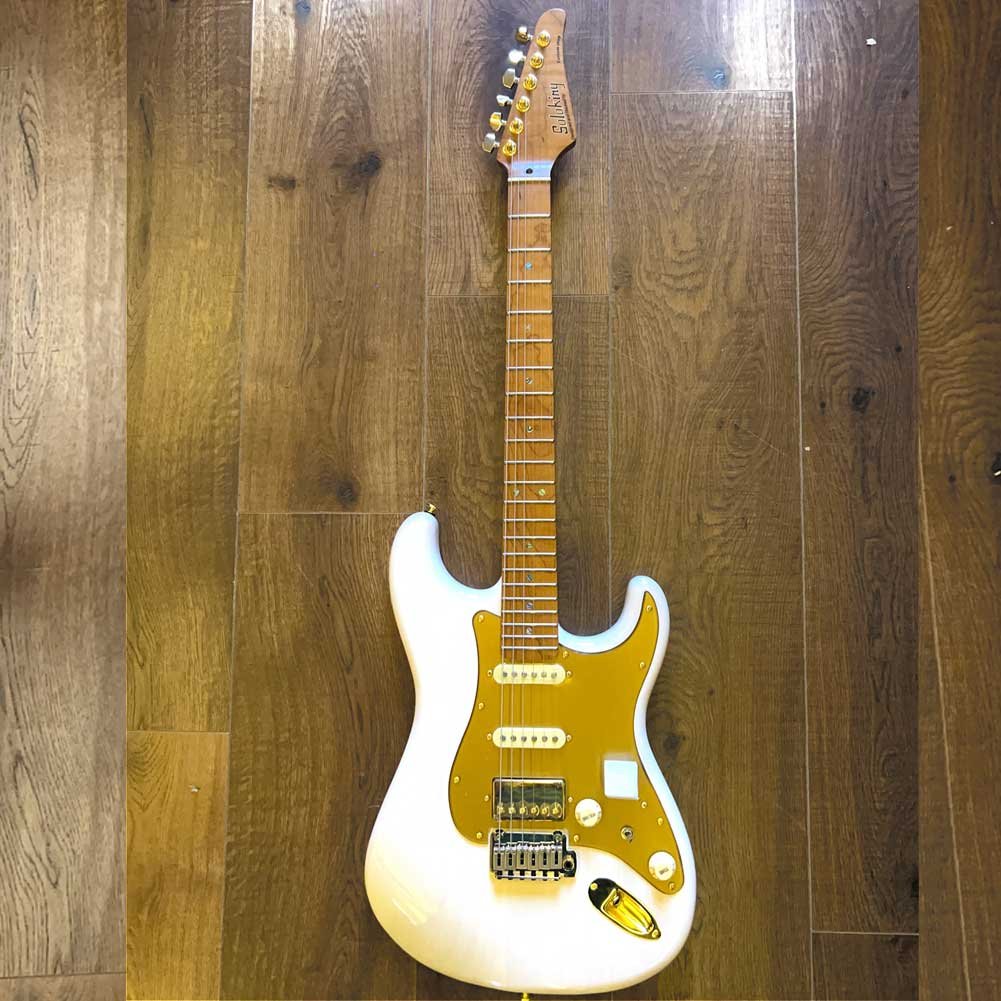 MS-1-Artisan-WB- White solid-body electric guitar with a gold pickguard resting on a wooden floor.